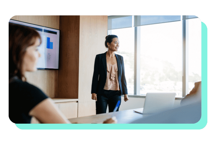 A woman stands confidently, presenting to colleagues in a conference room with a chart displayed on the screen.