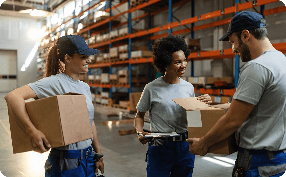 Three people in gray shirts and navy blue hats holding boxes in a warehouse.