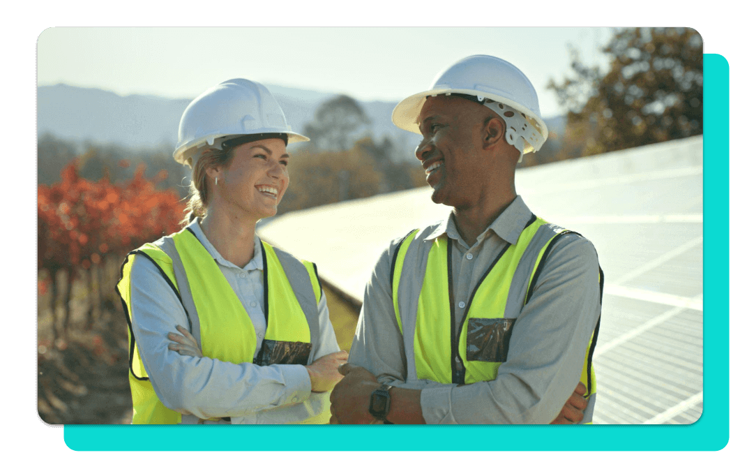 A man and woman talking. They are outside, wearing white hard hats and safety vests, in front of solar panels.